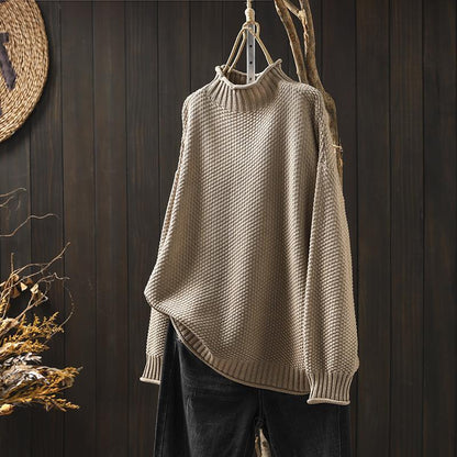 Maglione a collo alto beige in maglia a trama grossa, appeso su sfondo di legno scuro, accanto a decorazioni naturali. Moda autunnale elegante.