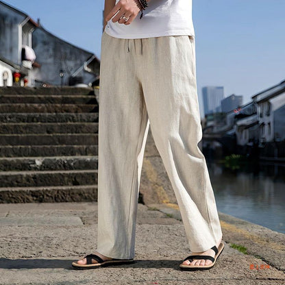 Uomo in pantaloni di lino beige e sandali neri su strada acciottolata vicino a canale, architettura tradizionale sullo sfondo.
