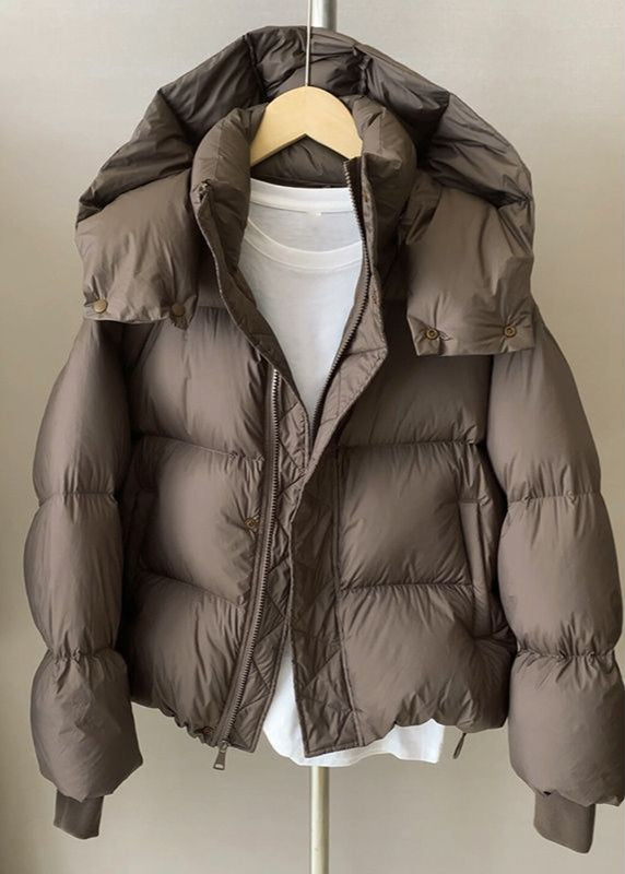 Giacca piumino marrone con cappuccio, appesa su gruccia, sopra maglietta bianca. Moda invernale, abbigliamento caldo, stile casual.