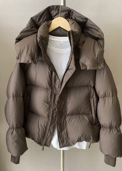 Giacca piumino marrone con cappuccio, appesa su gruccia, sopra maglietta bianca. Moda invernale, abbigliamento caldo, stile casual.