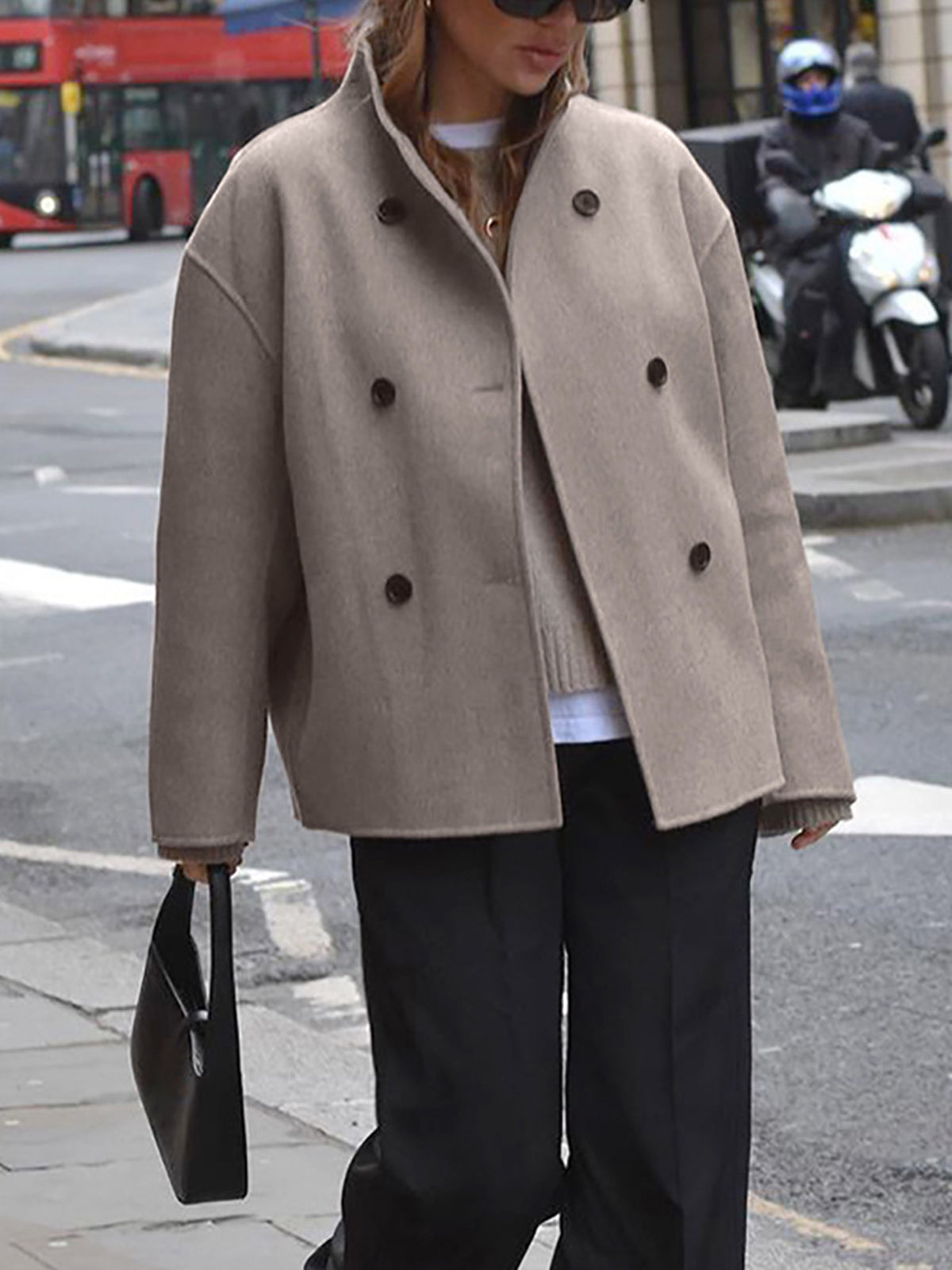 Donna con cappotto beige oversize e borsa nera, attraversa strada cittadina. Sfondo urbano con autobus rosso e scooter. Moda autunnale elegante.