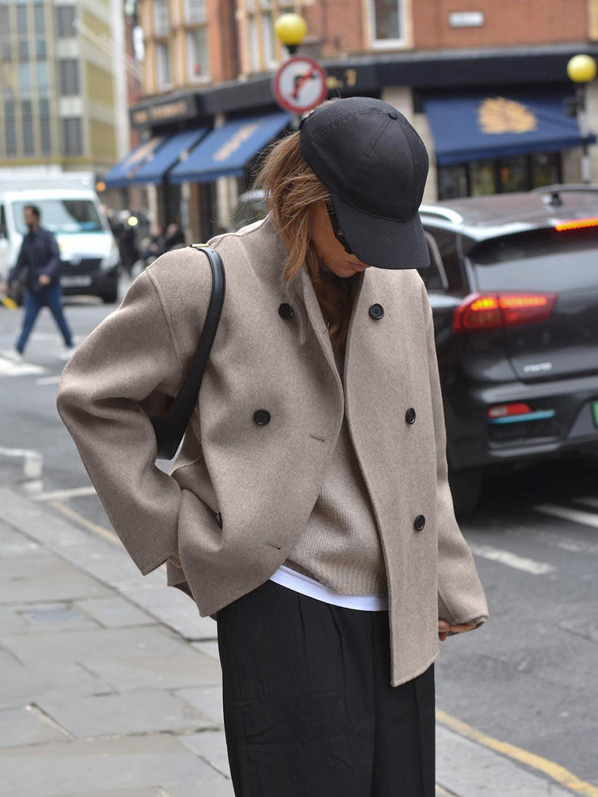 Donna con cappotto beige e cappello nero cammina in città. Moda autunnale, abbigliamento casual elegante, strada urbana, auto sullo sfondo.