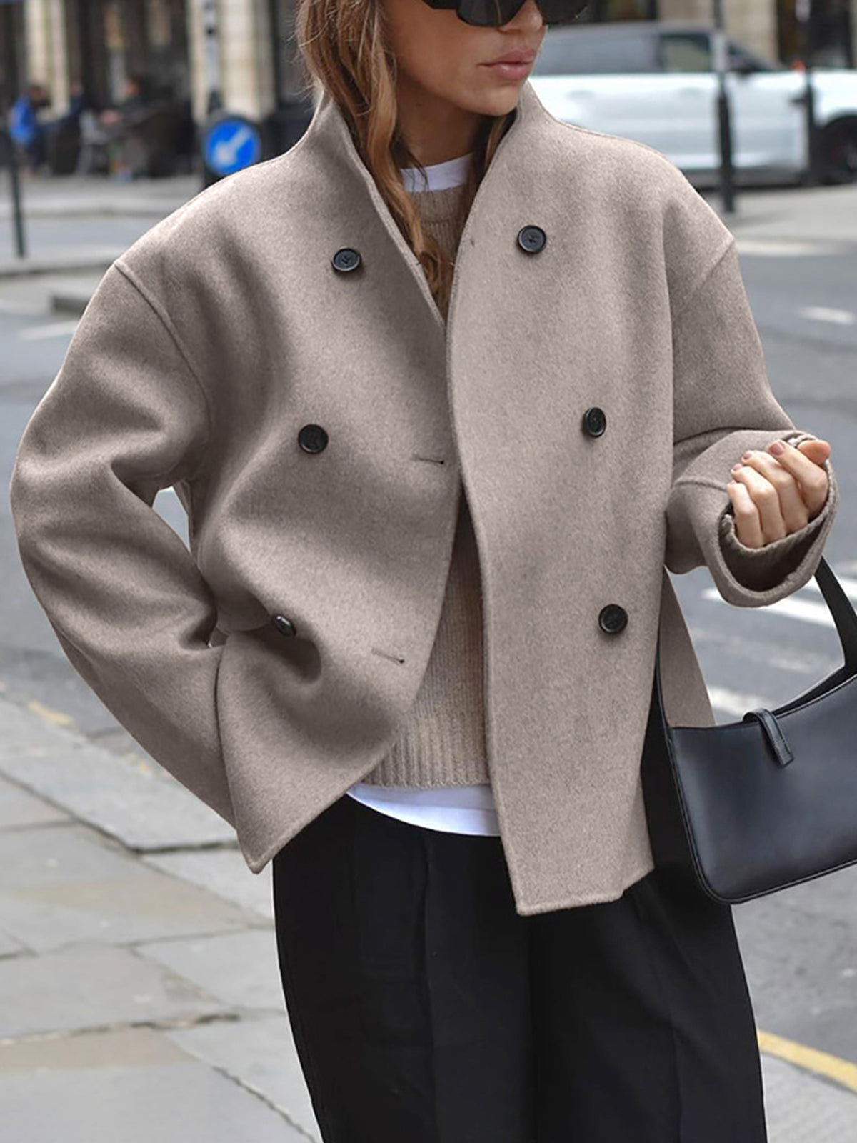 Donna con cappotto beige doppiopetto, occhiali da sole e borsa nera, cammina in città. Moda autunnale, stile urbano elegante.
