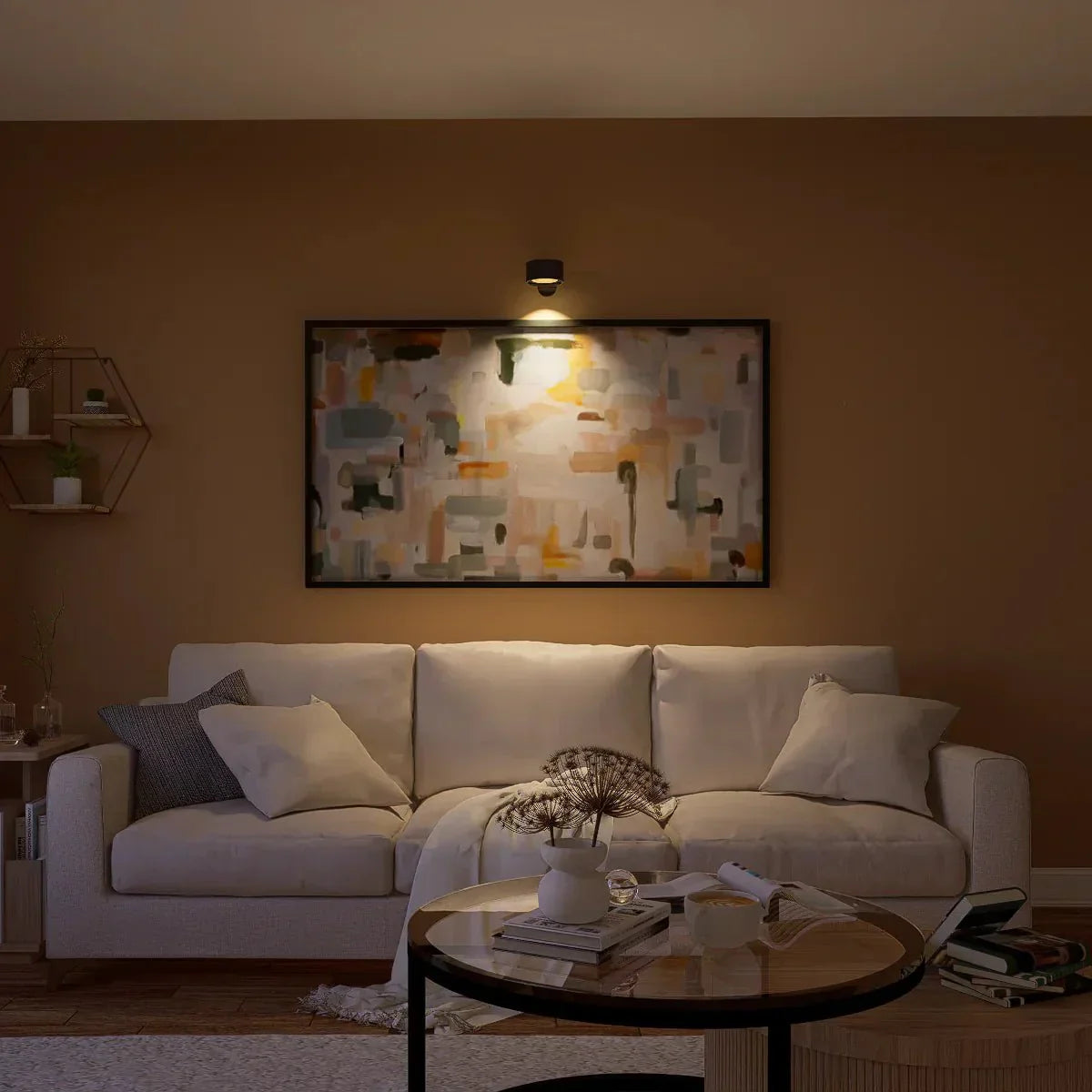 Divano beige in soggiorno moderno con pareti marroni, tavolino rotondo in legno, quadro astratto illuminato, cuscini decorativi e mensola esagonale.