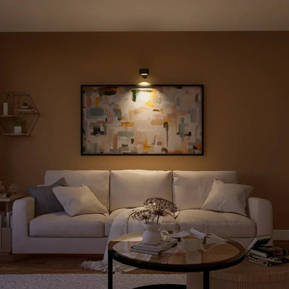 Divano beige in soggiorno moderno con pareti marroni, tavolino rotondo in legno, quadro astratto illuminato, cuscini decorativi e mensola esagonale.