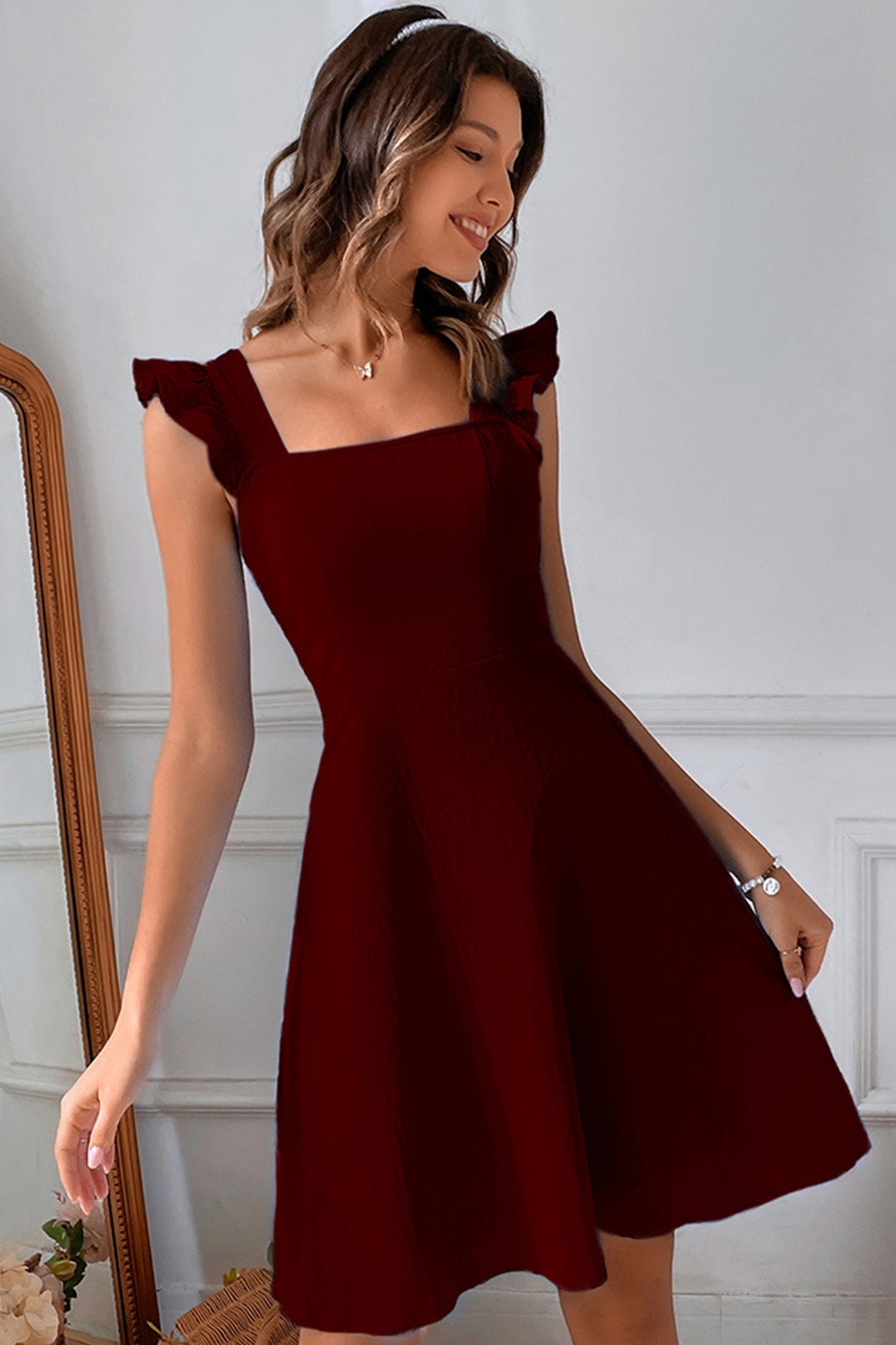 Donna in abito rosso bordeaux elegante, con spalline a volant, posa sorridente davanti a uno specchio. Moda femminile, abbigliamento da sera.