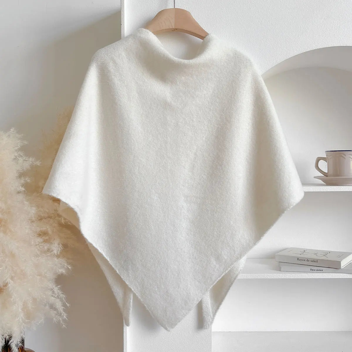 Poncho bianco in lana appeso su gruccia, design elegante e minimalista, ideale per moda autunno inverno, sfondo neutro con decorazioni leggere.