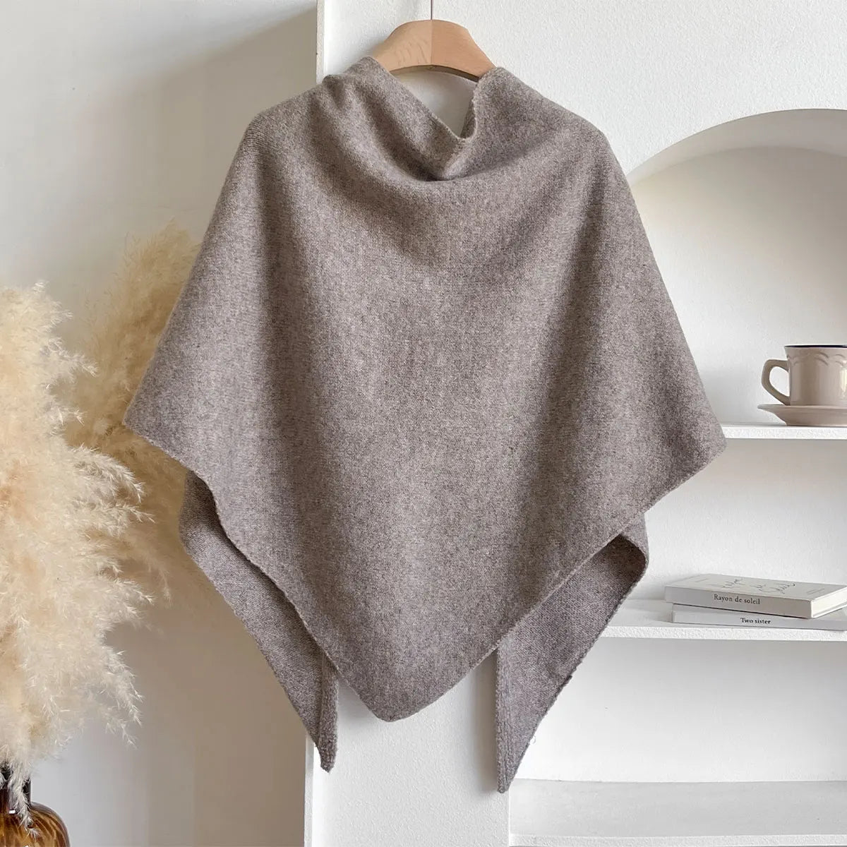 Poncho in lana grigia appeso su gruccia, sfondo minimalista con scaffale bianco e tazza. Elegante accessorio moda autunnale.