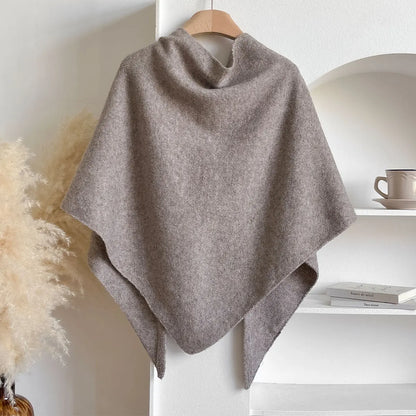 Poncho in lana grigia appeso su gruccia, sfondo minimalista con scaffale bianco e tazza. Elegante accessorio moda autunnale.