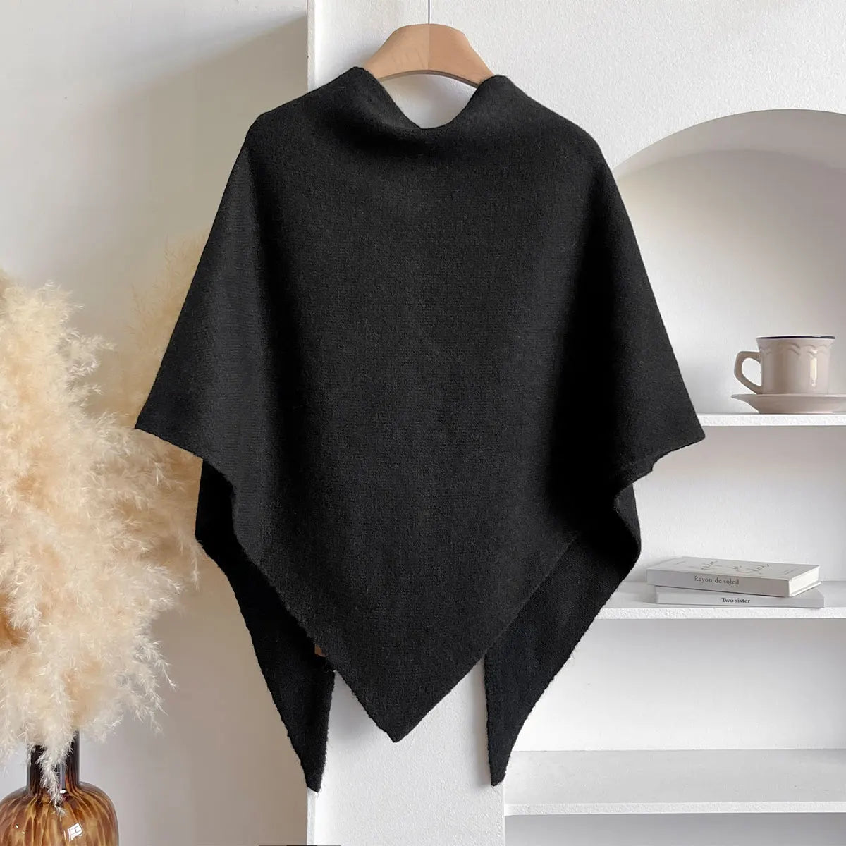 Poncho nero elegante appeso su gruccia, sfondo minimalista con scaffale bianco e tazza. Moda autunnale, accessorio caldo e versatile.
