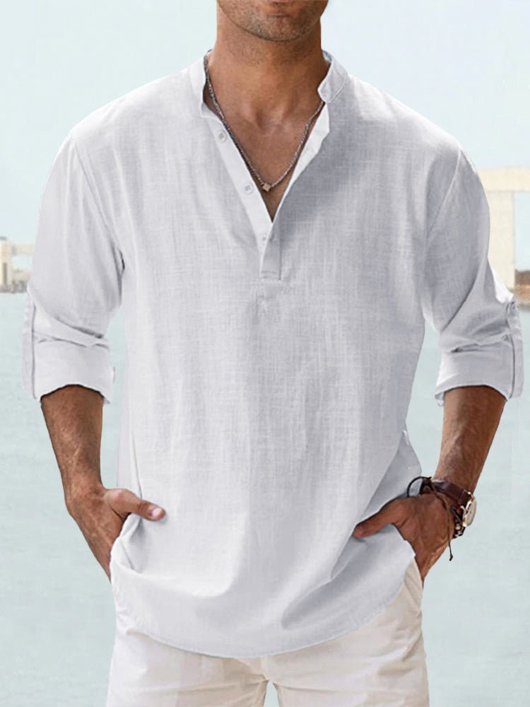 Camicia bianca da uomo in lino, maniche lunghe arrotolate, collo alla coreana, stile casual estivo. Sfondo marino sfocato.