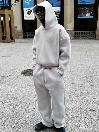 Uomo in tuta grigia con cappuccio e occhiali da sole, in piedi su marciapiede urbano. Moda streetwear, abbigliamento casual, stile moderno.