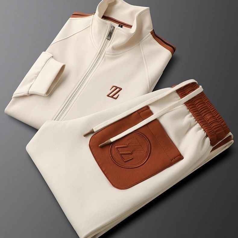 Tuta sportiva beige e marrone, con giacca a zip e pantaloni con tasca, su sfondo grigio. Abbigliamento casual elegante, moda uomo.