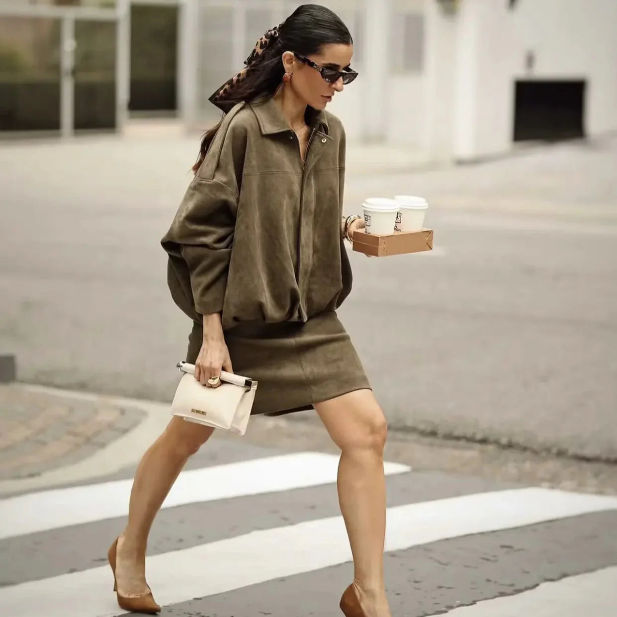 Donna elegante attraversa la strada con abito marrone, tacchi e occhiali da sole, portando caffè e borsa bianca. Moda urbana chic.