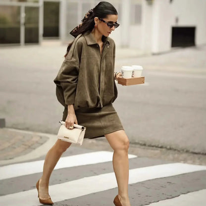 Donna elegante attraversa la strada con abito marrone, tacchi e occhiali da sole, portando caffè e borsa bianca. Moda urbana chic.