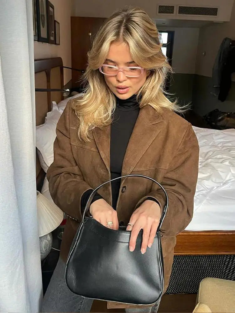 Donna bionda con occhiali rosa indossa giacca marrone e maglione nero, tiene borsa nera in camera da letto moderna. Moda autunnale elegante.