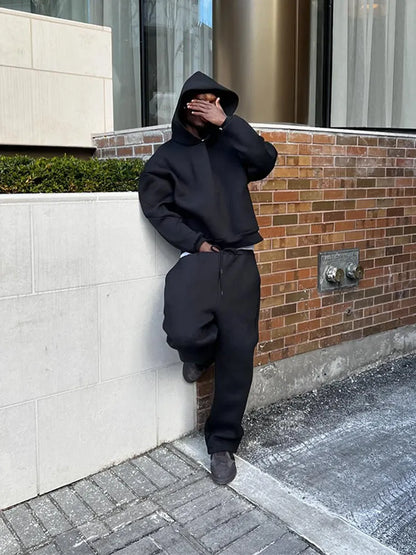 Uomo in abbigliamento sportivo nero con cappuccio, appoggiato a un muro di mattoni, copre il viso con la mano. Urban street style, moda casual.