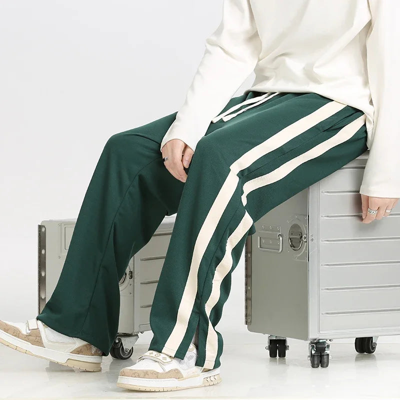 Pantaloni sportivi verdi con strisce bianche, indossati da una persona seduta su una valigia argentata. Scarpe da ginnastica beige visibili.