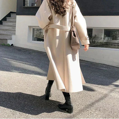 Donna con cappotto beige lungo, borsa a tracolla marrone, stivali neri, cammina su strada soleggiata. Moda autunnale elegante e casual.