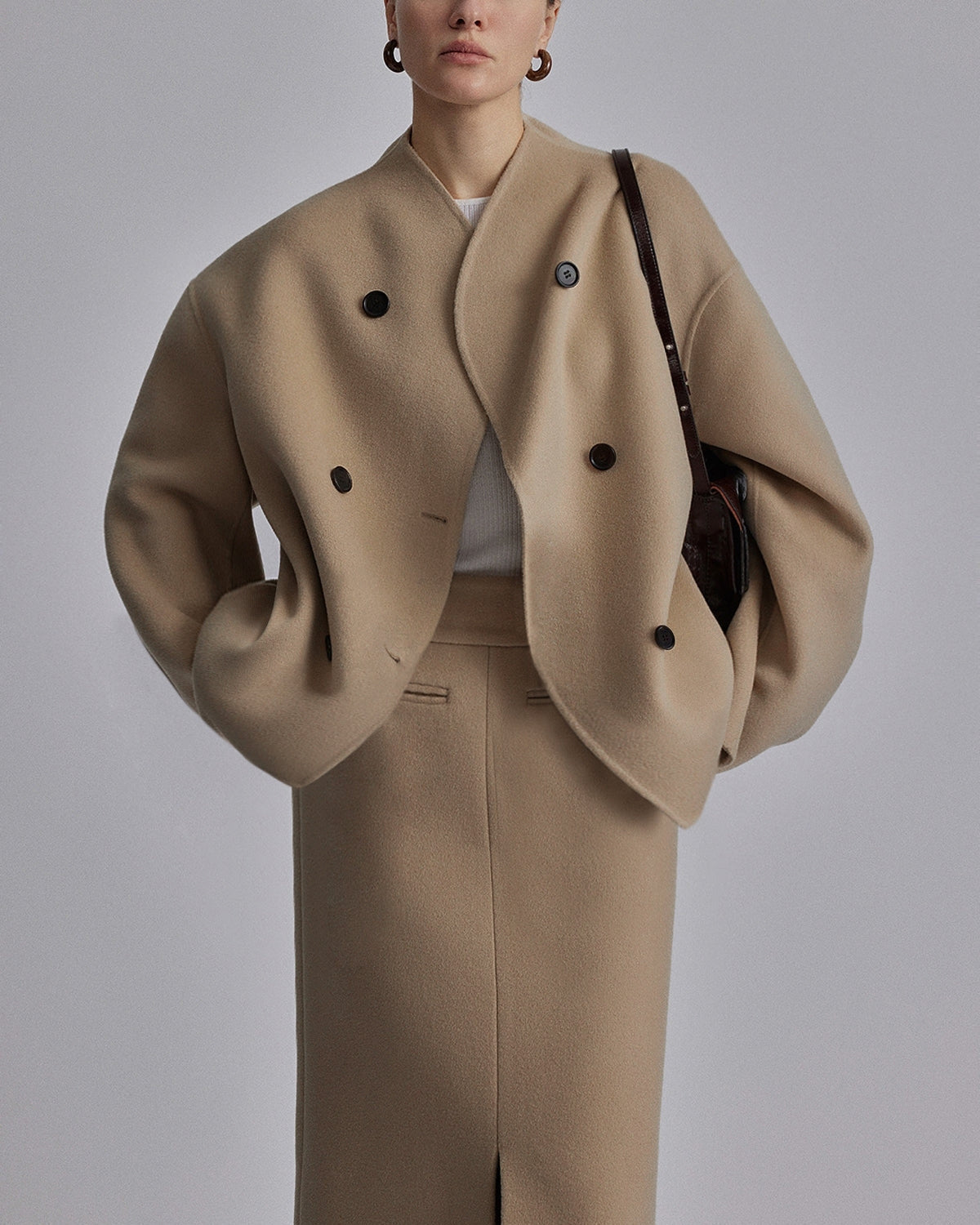 Donna con cappotto beige oversize e gonna coordinata, indossa orecchini a cerchio e borsa a tracolla marrone. Moda autunnale elegante e minimalista.