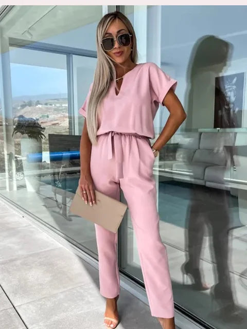 Donna in tuta rosa elegante, occhiali da sole, sandali beige, con clutch, in ambiente moderno con vetrate. Moda femminile estiva, stile chic.