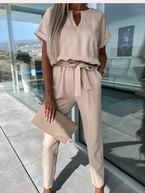 Donna in tuta beige elegante con cintura, pochette coordinata, capelli lunghi biondi, in terrazza moderna. Moda femminile, abbigliamento estivo.