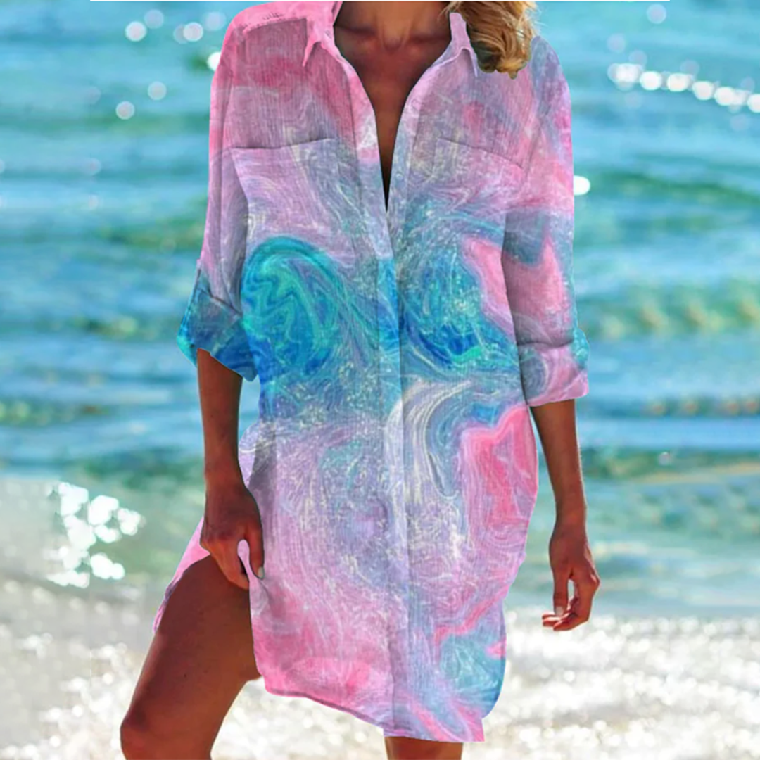 Donna in copricostume colorato con motivi astratti rosa e blu, cammina sulla spiaggia con mare sullo sfondo. Moda estiva, abbigliamento mare.