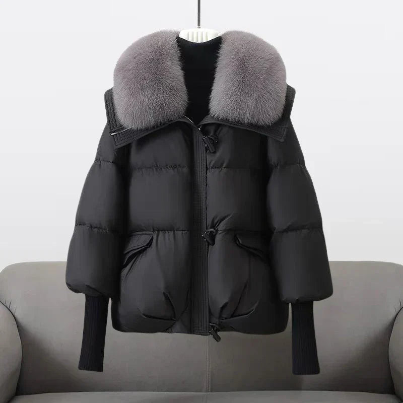 Giacca invernale nera imbottita con cappuccio e collo in pelliccia sintetica grigia, appesa su sfondo neutro. Moda donna, abbigliamento caldo.