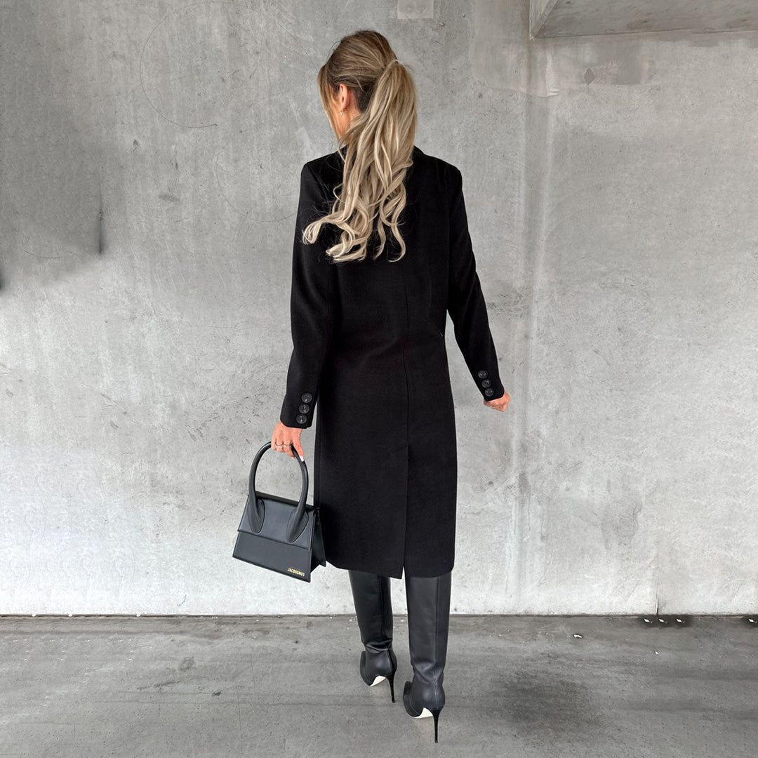 Donna di spalle con cappotto nero elegante, stivali alti e borsa a mano, su sfondo grigio. Moda autunnale, stile sofisticato.