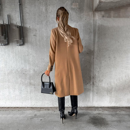 Donna di spalle con cappotto beige, stivali neri alti e borsa nera, sfondo grigio industriale. Moda autunno inverno, stile elegante urbano.