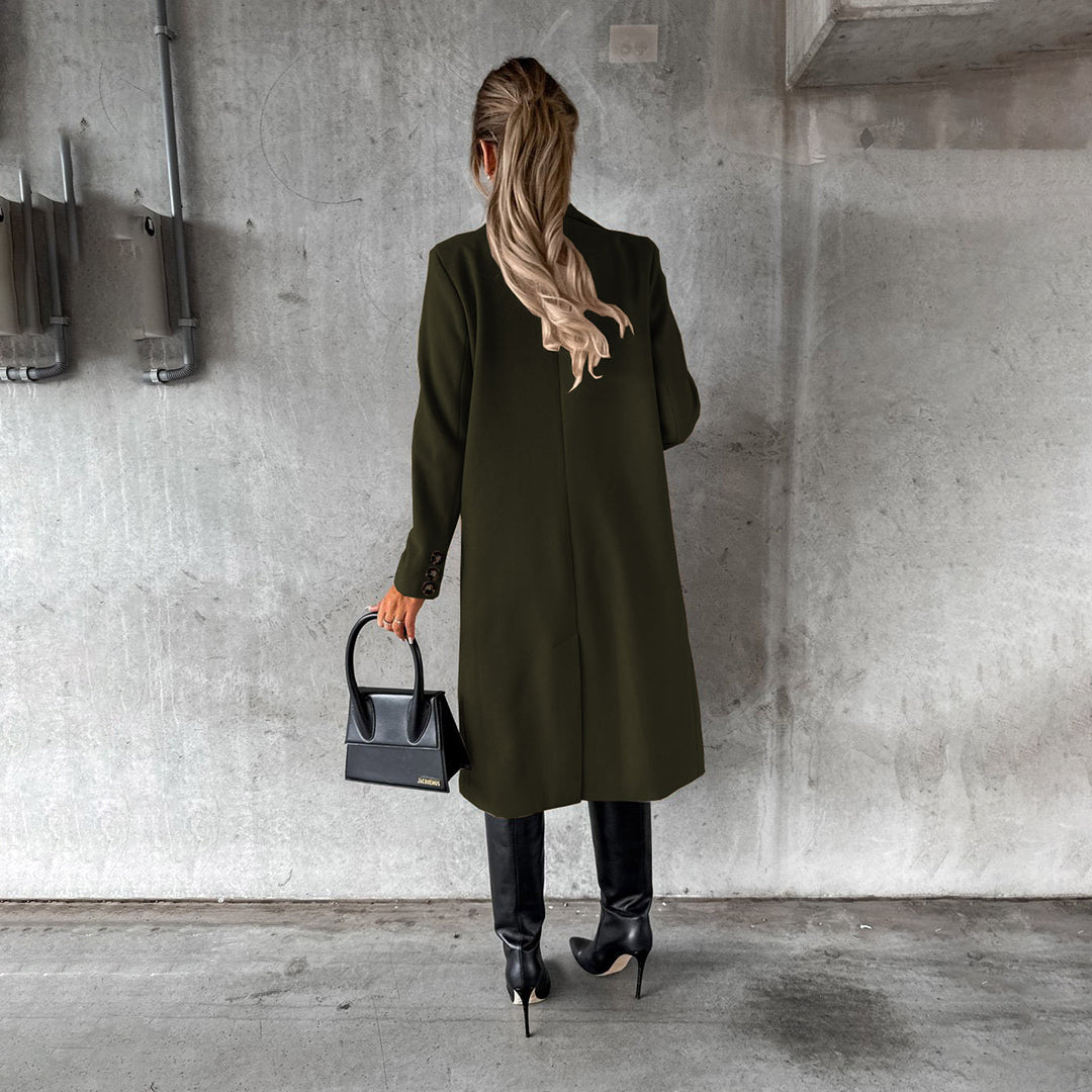 Donna di spalle con cappotto verde scuro, stivali neri e borsa a mano nera, su sfondo di cemento grigio. Moda autunnale elegante.