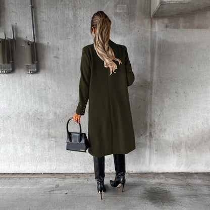 Donna di spalle con cappotto verde scuro, stivali neri e borsa a mano nera, su sfondo di cemento grigio. Moda autunnale elegante.