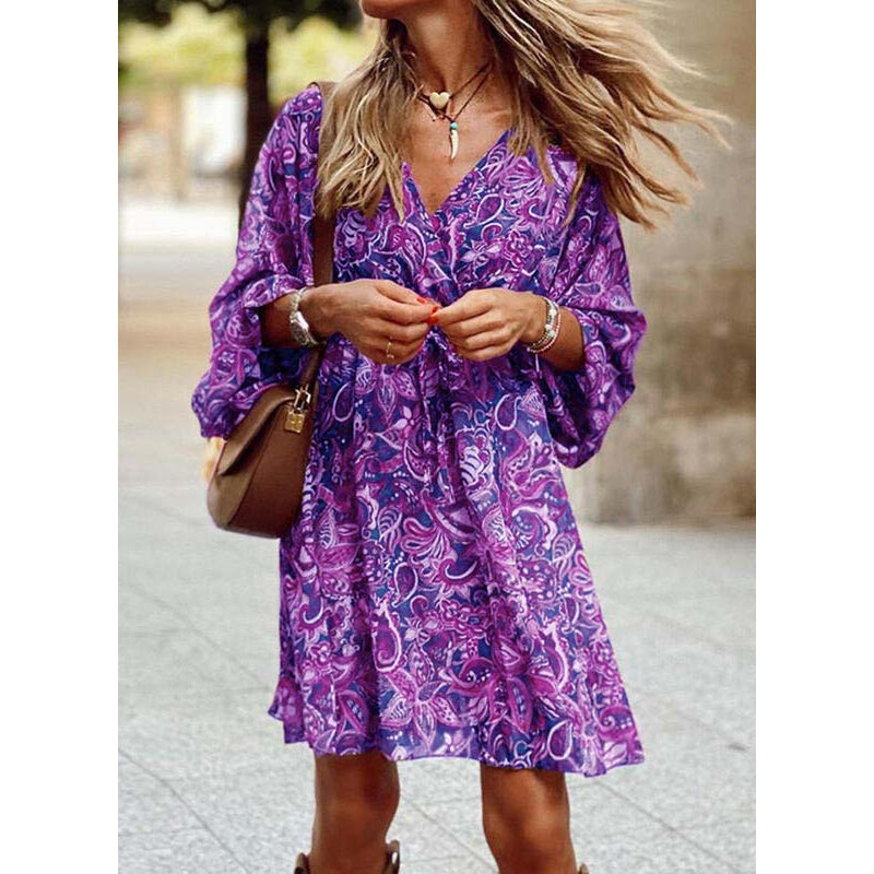 Donna con abito estivo viola a fantasia floreale, borsa marrone a tracolla, collana e bracciali, cammina all'aperto. Moda femminile casual.