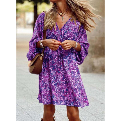 Donna con abito estivo viola a fantasia floreale, borsa marrone a tracolla, collana e bracciali, cammina all'aperto. Moda femminile casual.