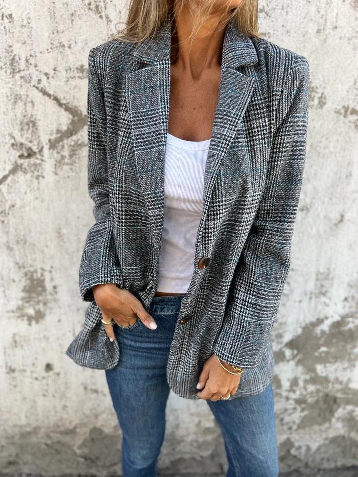 Donna con blazer a quadri grigio, top bianco e jeans, posa davanti a muro grezzo. Moda autunnale casual chic, stile urbano elegante.