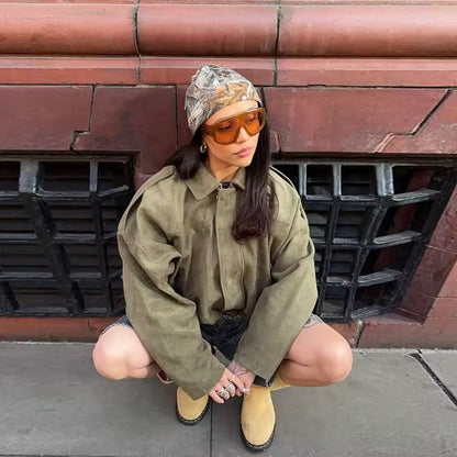 Donna con giacca oversize verde oliva, cappello mimetico e occhiali da sole arancioni, seduta su marciapiede urbano. Moda street style autunnale.