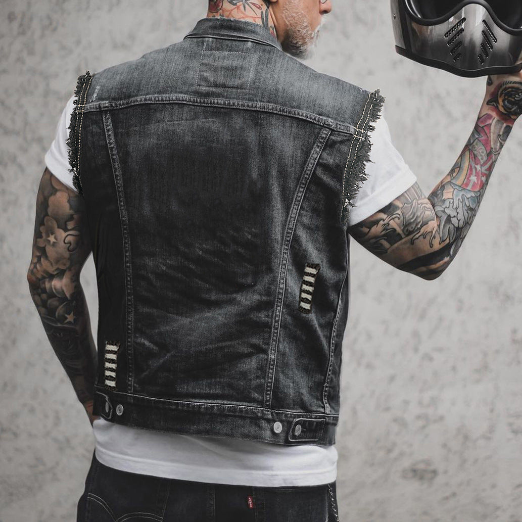 Giacca di jeans nera senza maniche, indossata da uomo tatuato, con maglietta bianca. Stile biker, moda urbana, dettagli strappati.