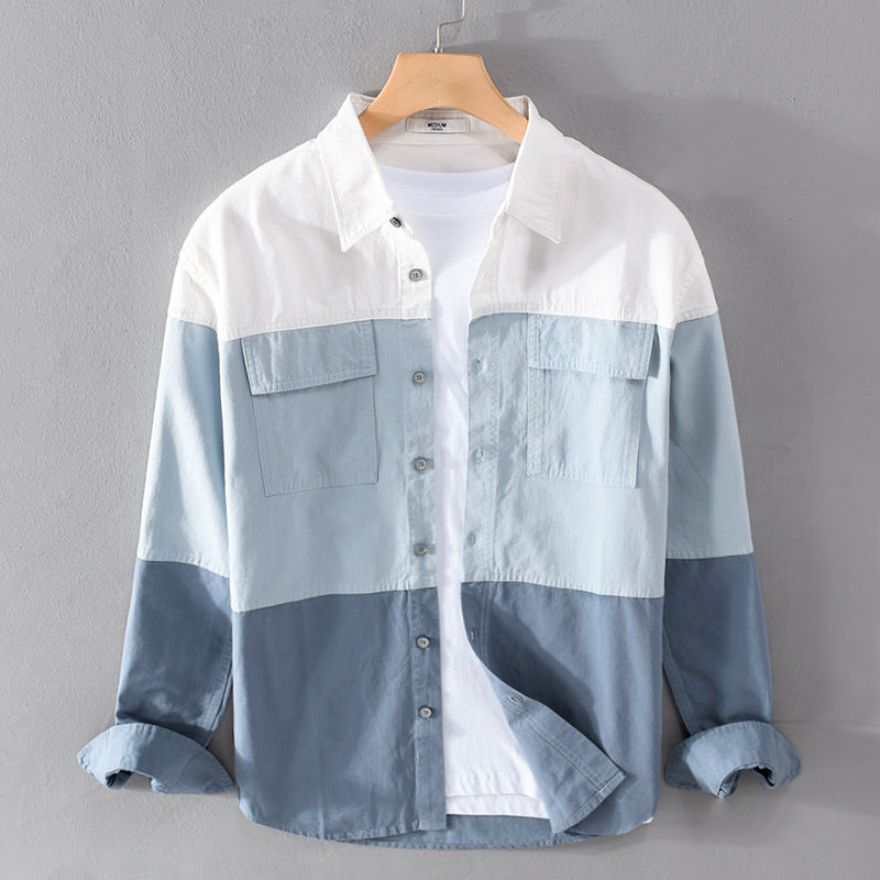 Camicia uomo a blocchi di colore blu e bianco, maniche lunghe, tasche frontali, stile casual, appesa su gruccia di legno, sfondo grigio.