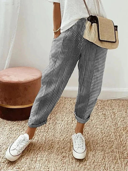 Donna con pantaloni a righe bianche e nere, borsa a tracolla in paglia, maglietta bianca e sneakers bianche su tappeto beige. Moda casual chic.