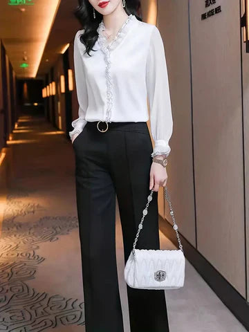 Donna elegante in camicia bianca con volant, pantaloni neri a vita alta, borsa bianca a tracolla, in corridoio moderno illuminato.