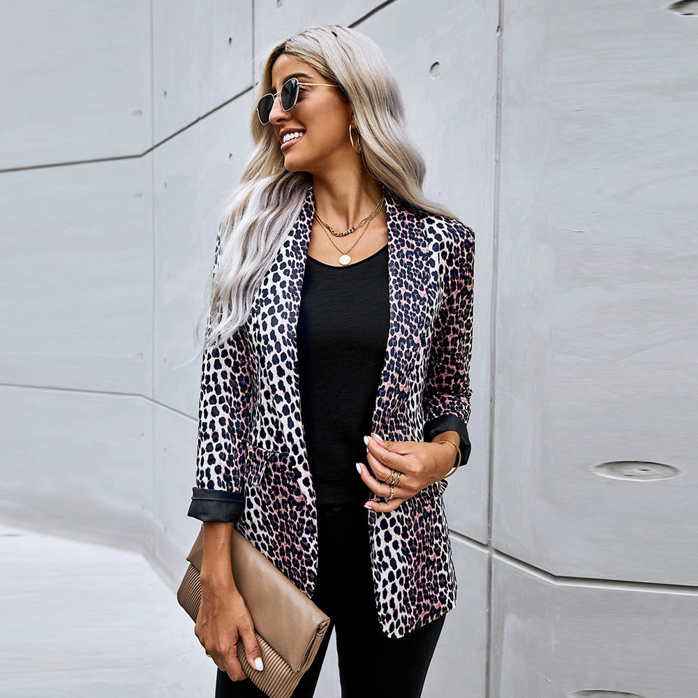 Donna con giacca leopardata, occhiali da sole e pochette beige, posa sorridente contro un muro moderno. Moda urbana, stile elegante, capelli biondi.