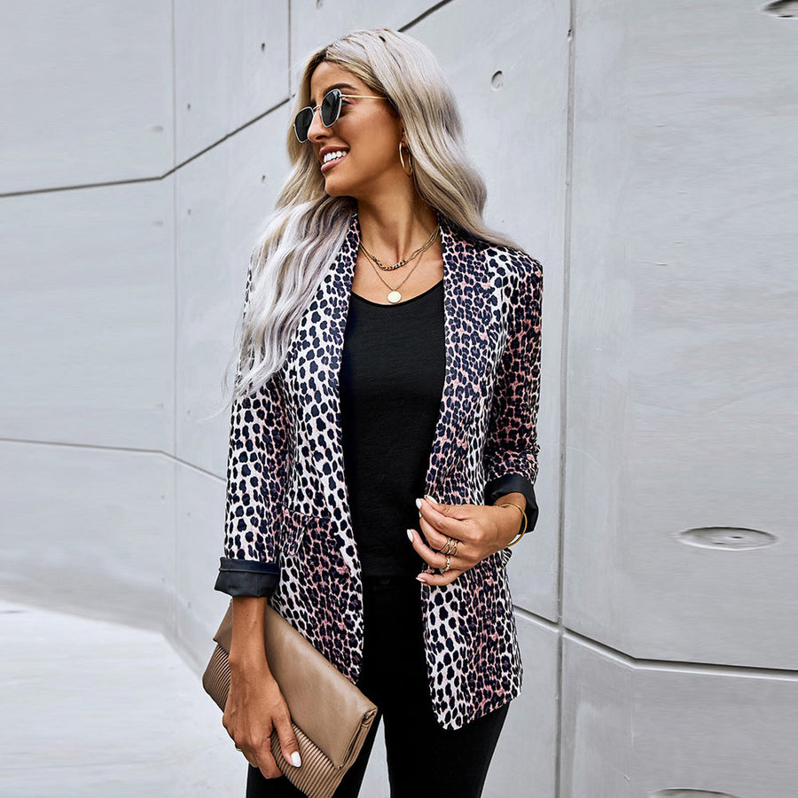 Donna con giacca leopardata, occhiali da sole e pochette beige, posa sorridente contro un muro moderno. Moda urbana, stile elegante, capelli biondi.