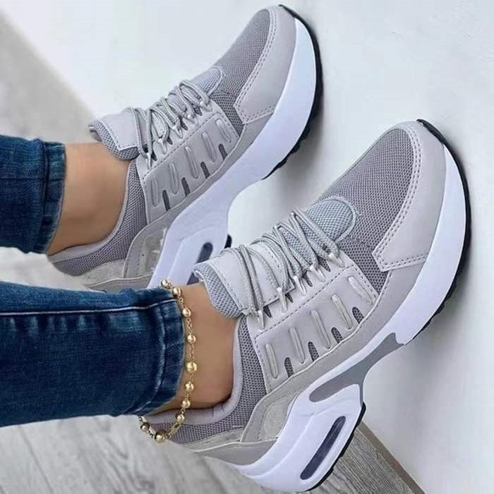 Scarpe da ginnastica grigie e bianche su pavimento in legno, indossate con jeans. Design moderno, suola ammortizzata, perfette per sport e tempo libero.
