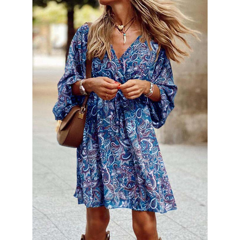 Donna con abito estivo blu a fantasia paisley, borsa a tracolla marrone, collana e bracciali, capelli mossi al vento, in strada cittadina.