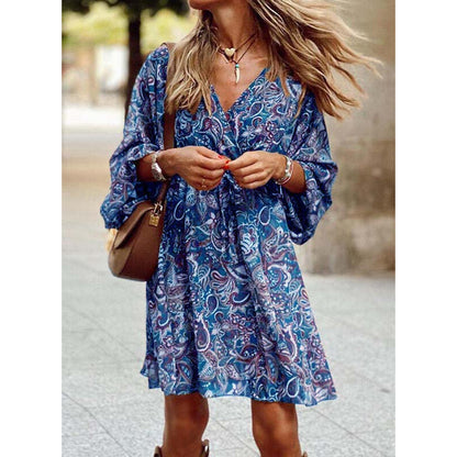 Donna con abito estivo blu a fantasia paisley, borsa a tracolla marrone, collana e bracciali, capelli mossi al vento, in strada cittadina.