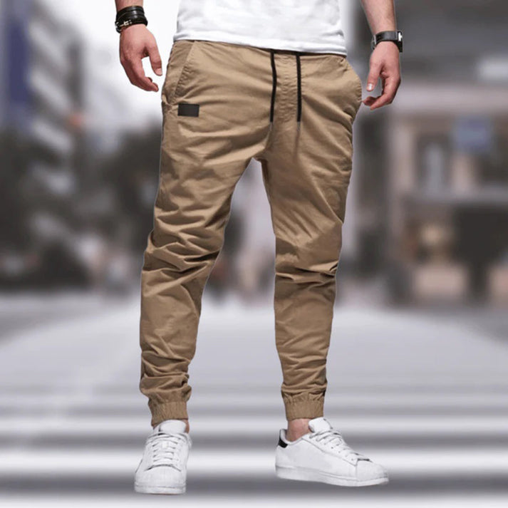 Uomo con pantaloni jogger beige e scarpe da ginnastica bianche su sfondo urbano sfocato. Moda casual maschile, streetwear moderno.
