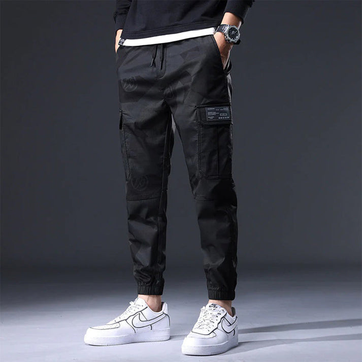 Pantaloni cargo neri da uomo, stile casual, con tasche multiple, abbinati a scarpe da ginnastica bianche. Moda streetwear moderna.