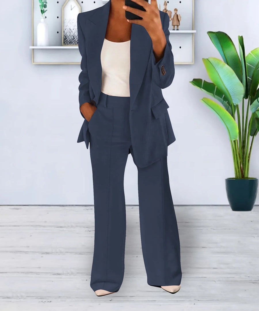 Donna in elegante completo blu scuro, giacca e pantaloni, con top bianco, in un ambiente moderno con pianta verde e scaffale decorativo.