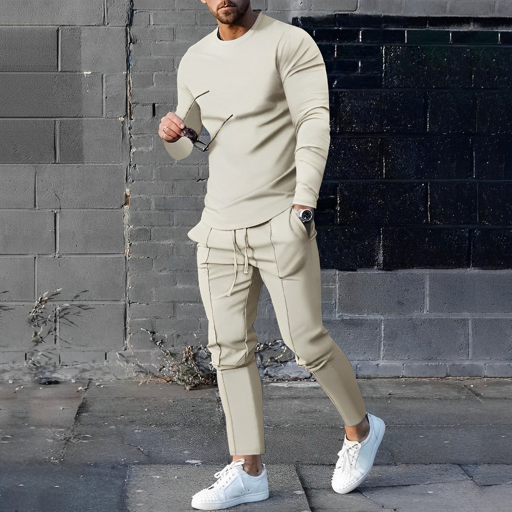 Uomo in tuta beige alla moda, con occhiali da sole e orologio, posa su sfondo urbano. Scarpe bianche, look casual ed elegante.