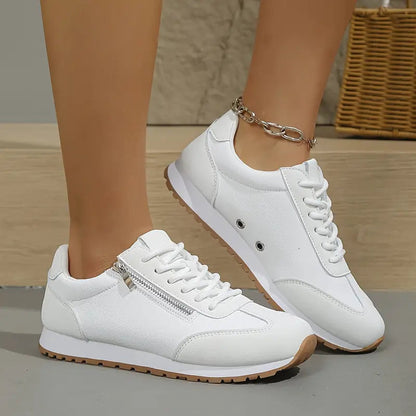 Scarpe da ginnastica bianche da donna con cerniera laterale, suola in gomma marrone, indossate su pavimento in legno. Accessorio cavigliera in metallo.